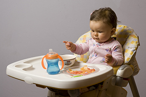 Un bébé assis dans une chaise haute avec une tasse à bec, tentant d'atteindre des amuse-gueule.